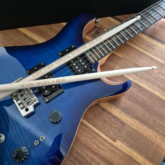 A blue electric guitar with two white drumsticks resting across it on a wooden floor. The drumsticks are engraved with 'UNITE UNION NATIONAL SECRETARY 10/20/20' and 'NEVER MISSES A BEAT'.
