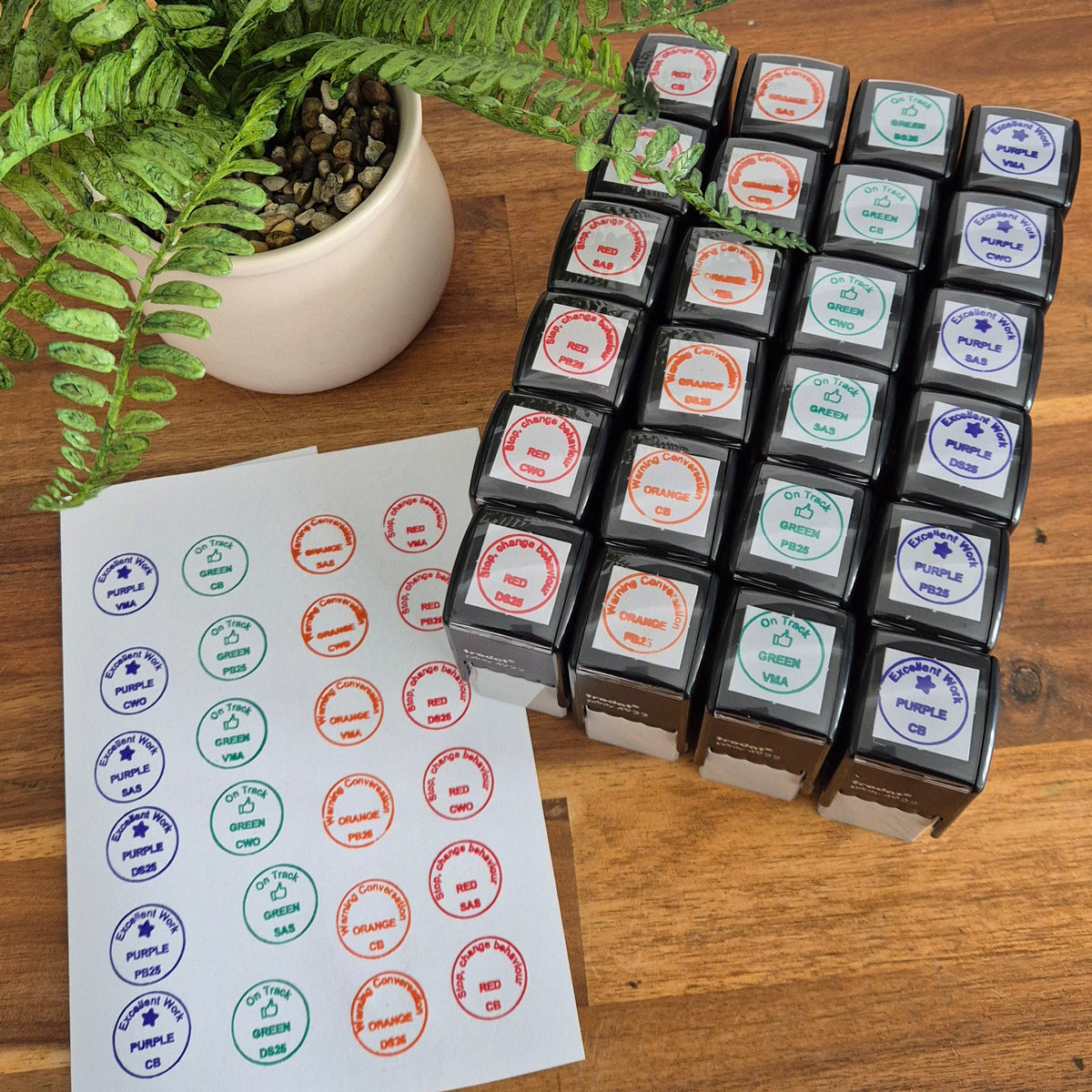A set of small square ink pads or stamp blocks arranged on a wooden table next to a sheet of circular printed stamps, with a potted fern in the background.