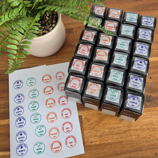 A set of small square ink pads or stamp blocks arranged on a wooden table next to a sheet of circular printed stamps, with a potted fern in the background.