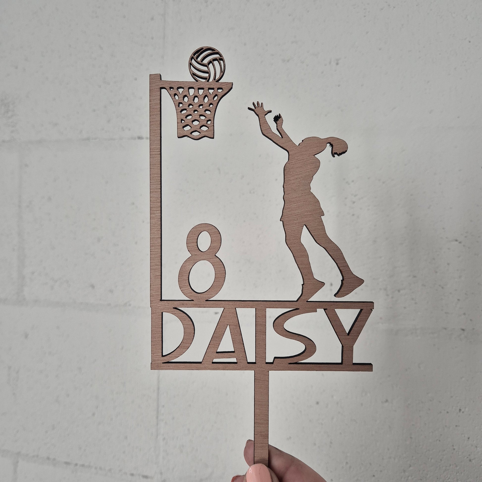Decorative wooden cake topper with a netball player and Daisy branding on a light gray background.