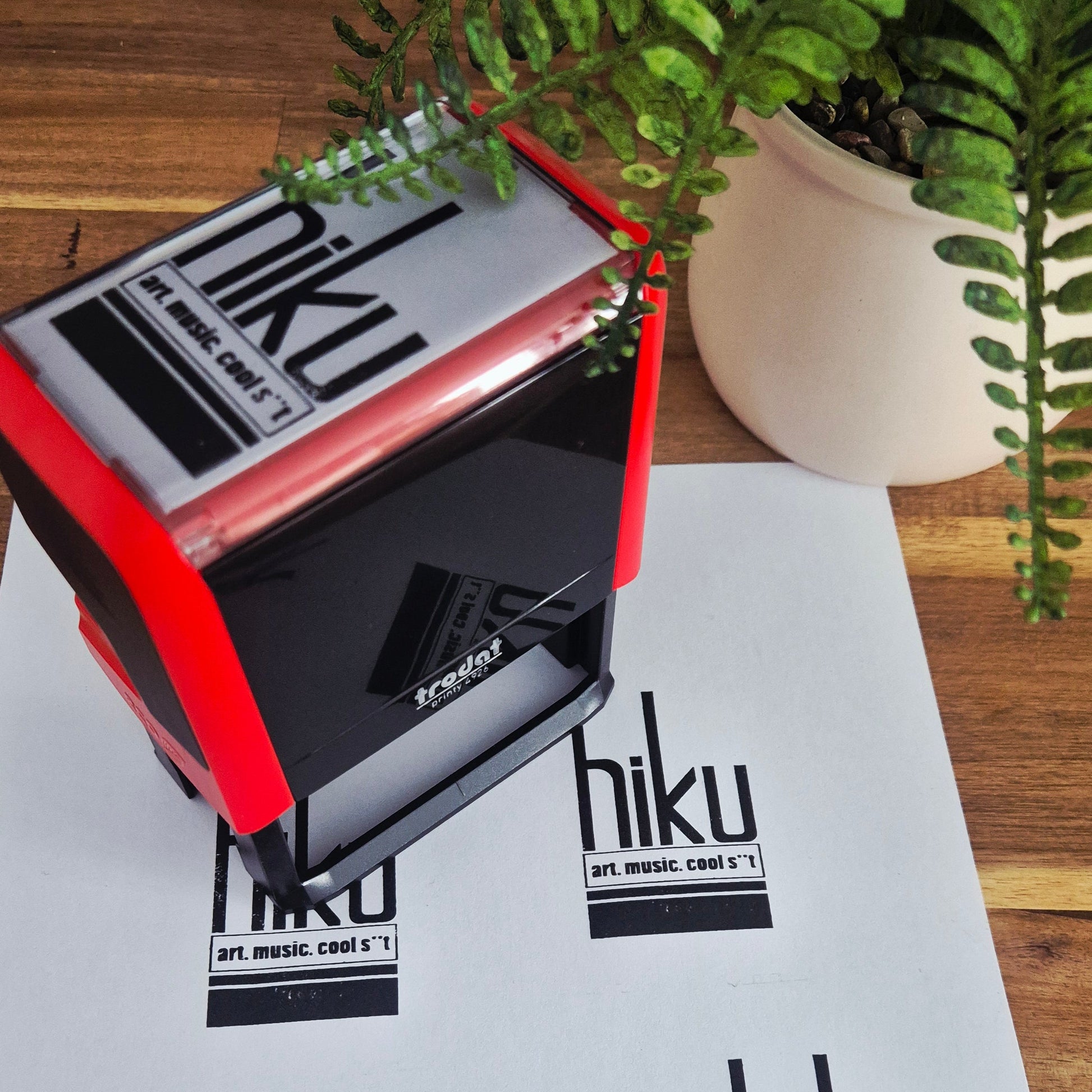 A red and black handheld rubber stamp with the word 'hiku' is shown pressing onto a sheet of white paper that already has matching stamped logos. A potted green fern sits on a wooden table in the background.