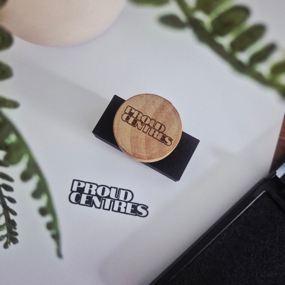 Wooden engraved handle with 'Proud Centres' branding on black 3D Printed Stamp block and a stamped impression on white paper with green leaves.