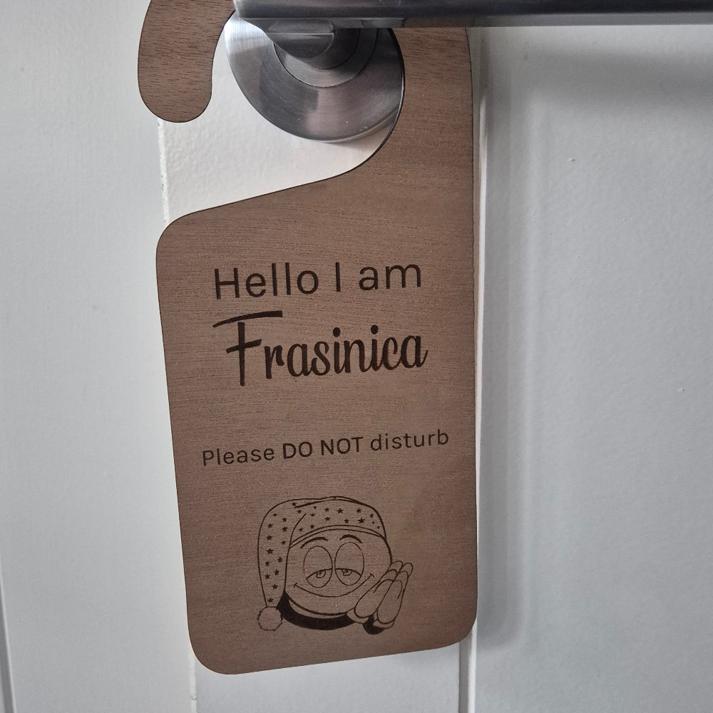 A wooden hanger tag with a metal hook, printed with 'Hello I am Frasinetea Please DO NOT disturb' and a small illustration, hanging on a white door.