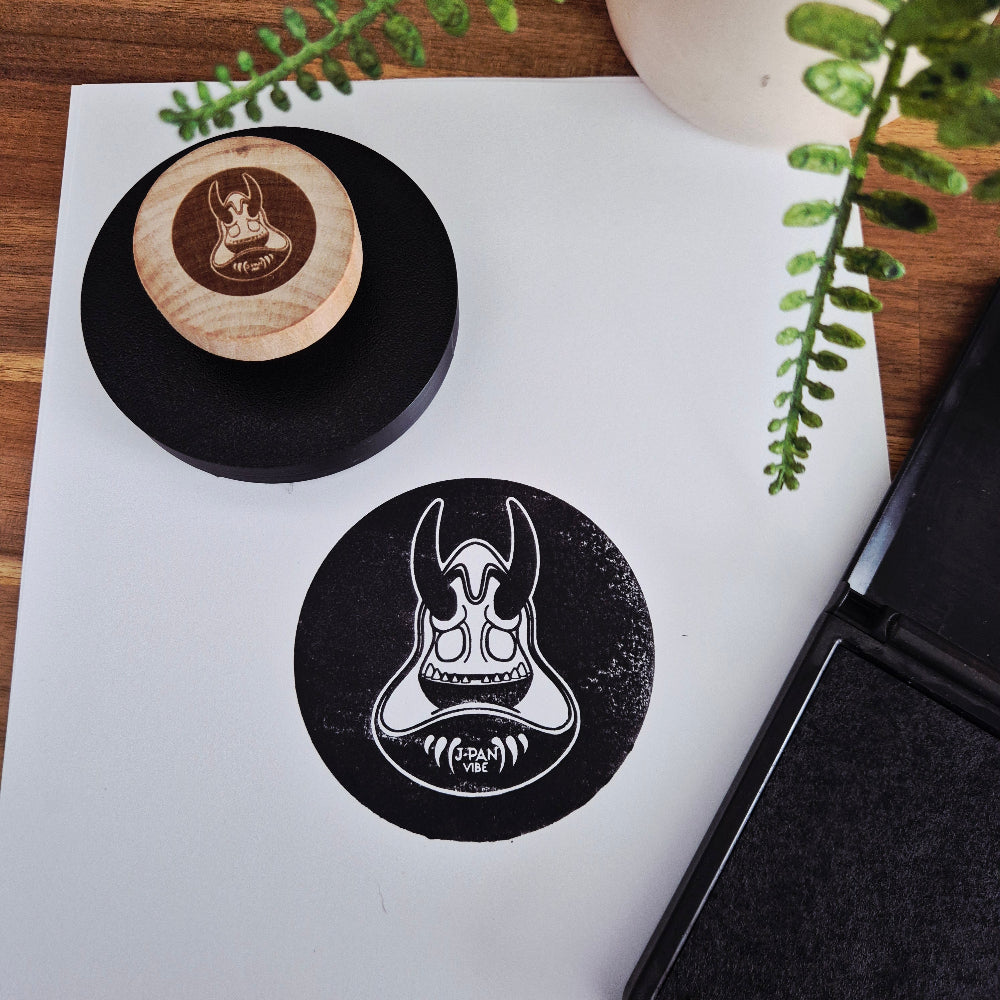Large circular 3D Printed Handheld stamp with an engraved wooden handle stamped onto white paper with Japan Vibe Logo.  