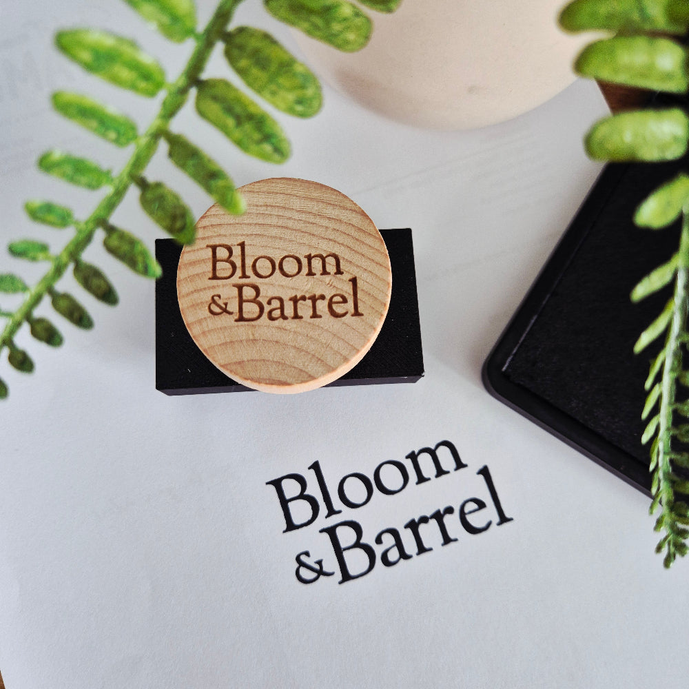 Medium Handheld Stamp with 3D Printed stamp block and engraved wooden handle with 'Bloom & Barrel' branding on a white surface with green leaves.