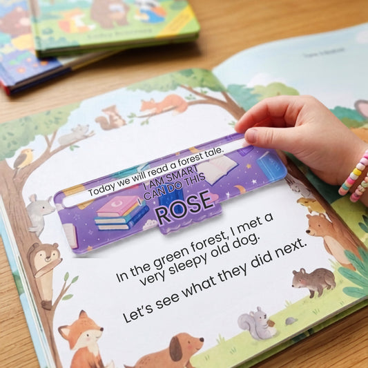 A child's hand with colorful beaded bracelets holds a purple plastic bookmark over an open children's picture book on a wooden table.