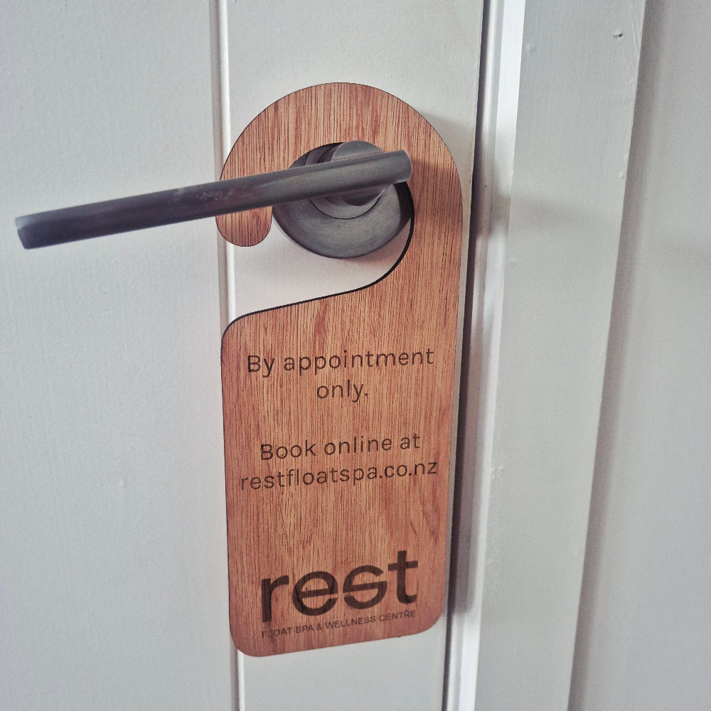 A wooden doormat sign with a curved hook designed for a door handle, featuring the brand name 'reet' and booking information printed on it, shown hanging on a white door.