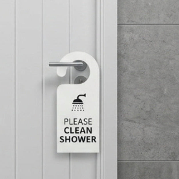 A white bathroom door with a metal handle has a white hanging sign that reads 'PLEASE CLEAN SHOWER' with a shower icon. A gray tiled wall and a small 'SHOWER ROOM 1' plaque are visible.