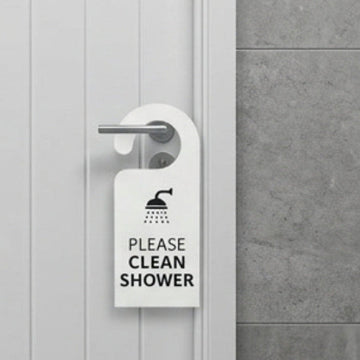 A white bathroom door with a metal handle has a white hanging sign that reads 'PLEASE CLEAN SHOWER' with a shower icon. A gray tiled wall and a small 'SHOWER ROOM 1' plaque are visible.
