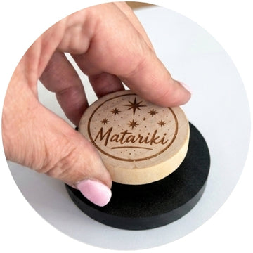 A close-up of a hand pressing down on a custom circular wooden stamp with the name 'Matariki' and stars engraved on it, resting on a black circular base.