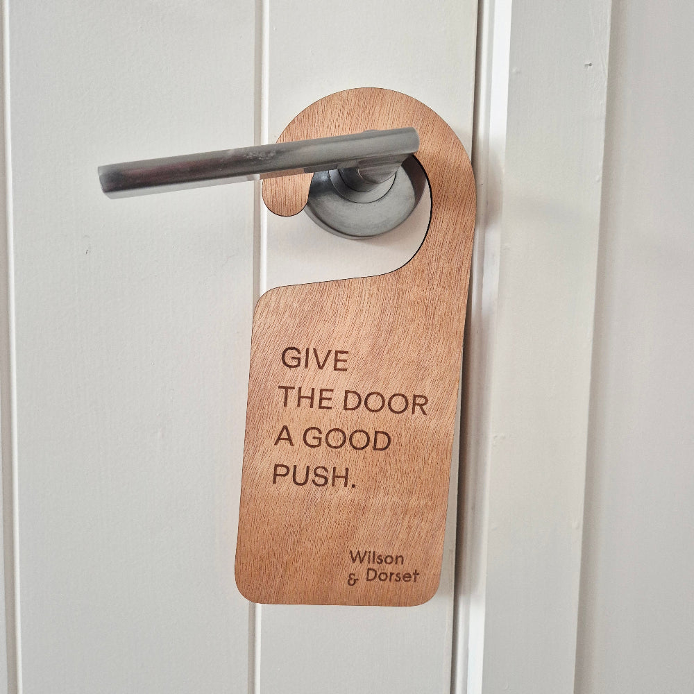 A wooden do-not-disturb door hanger with engraved text reading 'GIVE THE DOOR A GOOD PUSH.' and 'Wilton & Davenport' on the right, shown hanging from a silver metal door handle on a white paneled door.