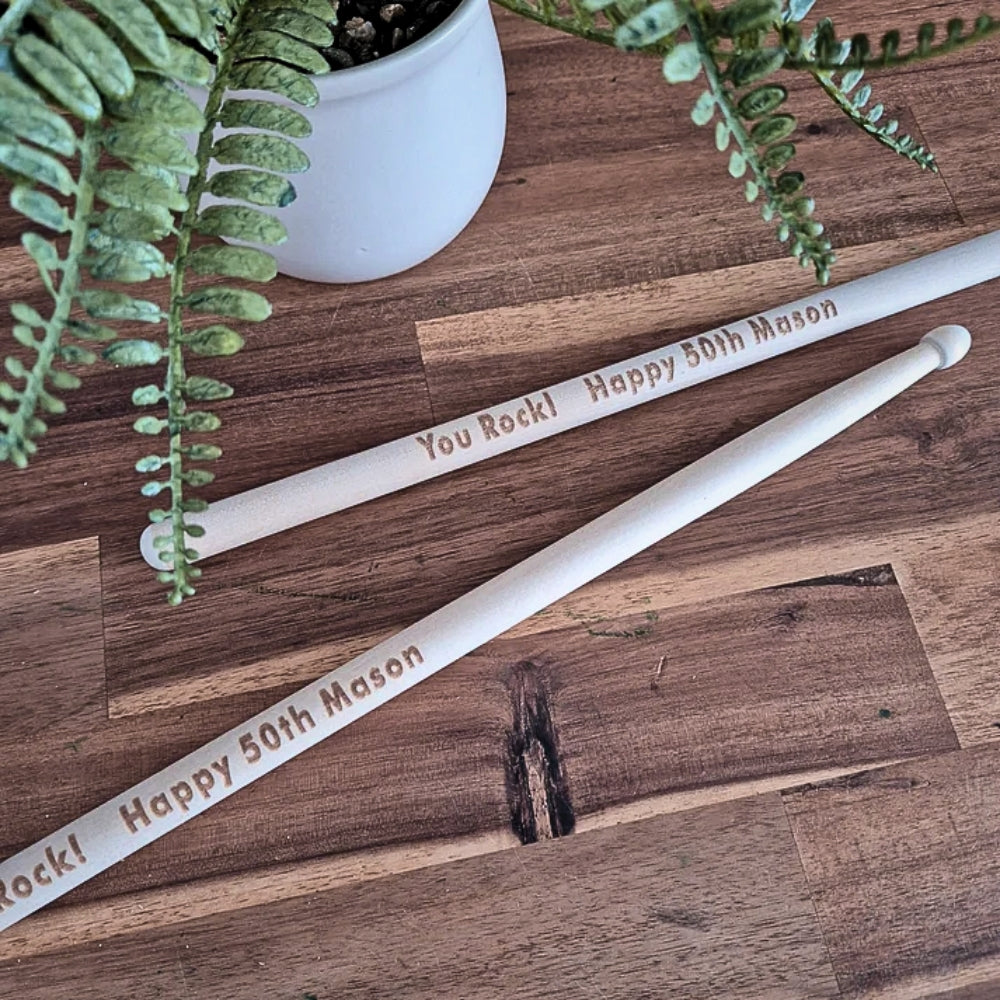 Two white wooden birthday candles engraved with 'You Rock! Happy 50th Mason' in brown lettering, arranged on a rustic wooden tabletop next to a potted fern.