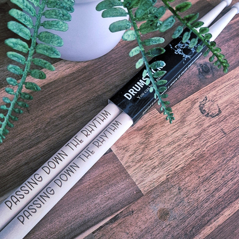 A pair of drumsticks with engraved text 'PASSING DOWN THE RHYTHM' resting on a wooden surface next to a black drumstick tip sleeve labeled 'DRUMSTICKS'. Green plant leaves and a white vase are visible in the background.