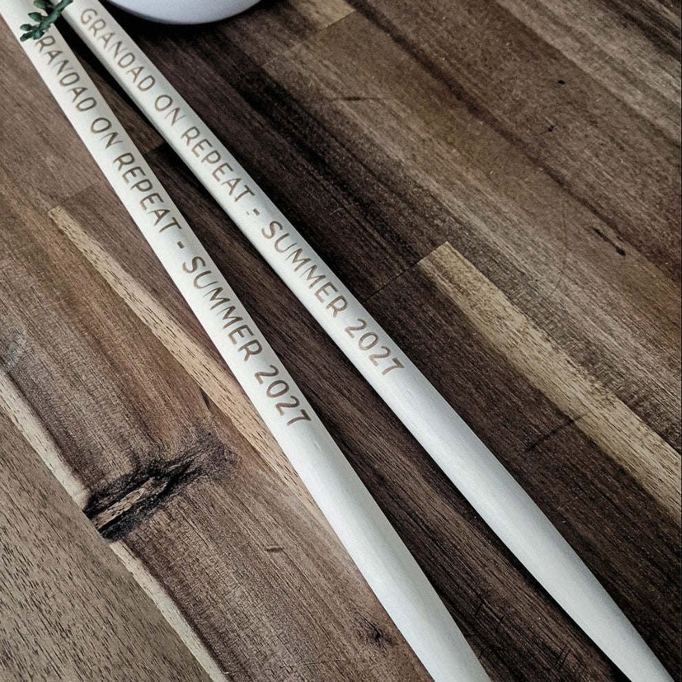 Two white wooden drumsticks engraved with 'GRANDAD ON REPEAT - SUMMER 2027' in brown lettering, placed diagonally on a dark wood background.