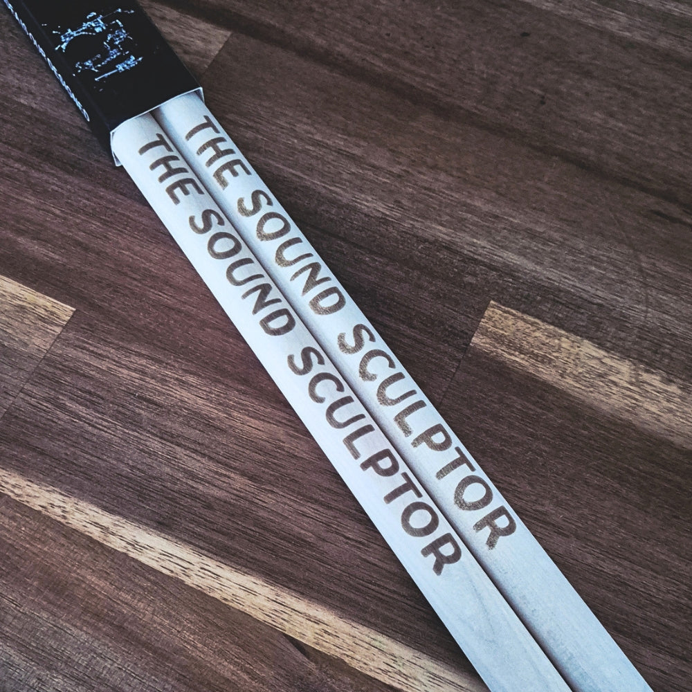A pair of white drumsticks with 'THE SOUND SCULPTOR' printed in brown on the sticks, resting on a dark wood plank surface.