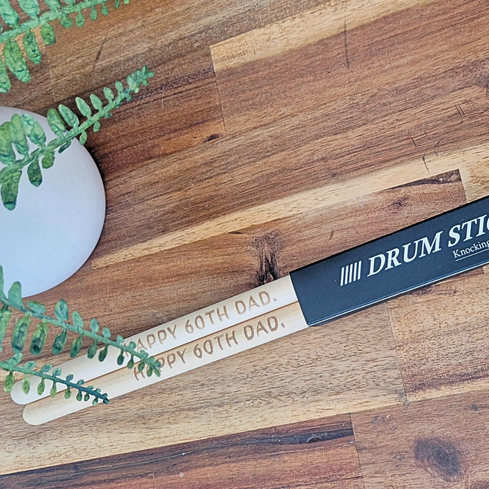 A pair of engraved drum sticks with a black handle, placed on a wooden surface next to a white vase with green leaves. The sticks are engraved with 'HAPPY 60TH DAD.