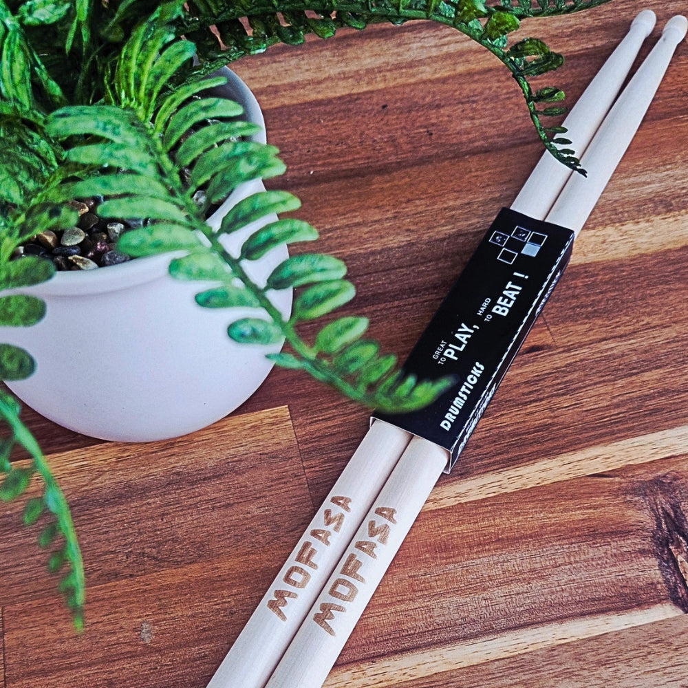 A pair of white drumsticks wrapped in a black band labeled 'PLAY BEAT I DRUMSTICKS' and engraved with 'MUFASA' and 'MUFASA', placed on a wooden surface next to a potted green plant.