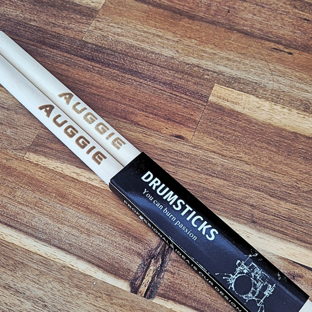 A pair of drumsticks with 'AUGGIE' printed on them, resting on a black sleeve with the text 'DRUMSTICKS You can burn passion' on a wooden surface.