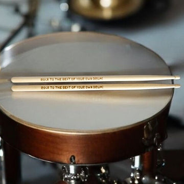 A pair of wooden drumsticks resting on the drumhead of a drum, printed with the text 'ROCK TO THE BEST OF YOUR OWN DRUM!'.