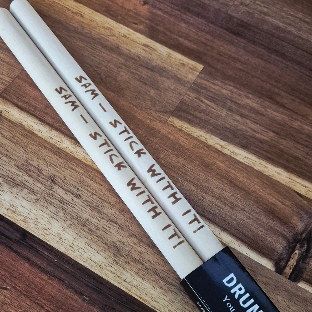 A pair of drumsticks with engraved text reading 'SAM - STICK WITH IT!' resting on a wooden surface. One end of the sticks is black with white text that reads 'DRUM You'.