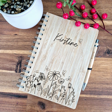 A bamboo notebook with a natural finish, featuring personalized engraving of floral designs and the name 'Kirstine'. It is accompanied by a pen and displayed on a wooden surface.