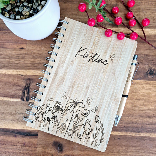 A bamboo notebook with a natural finish, featuring personalized engraving of floral designs and the name 'Kirstine'. It is accompanied by a pen and displayed on a wooden surface.