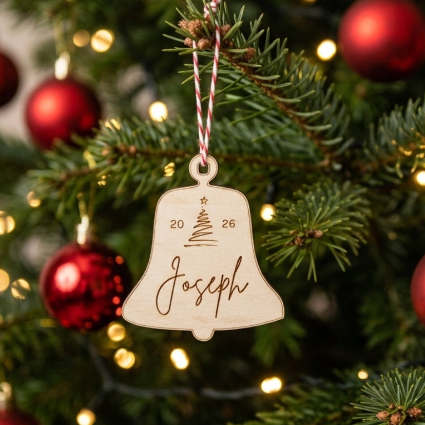 A personalized wooden Christmas bell ornament engraved with the name 'Joseph' and the dates '20' and '26', hanging from a red and white striped ribbon on a decorated Christmas tree with red ornaments and warm lights.