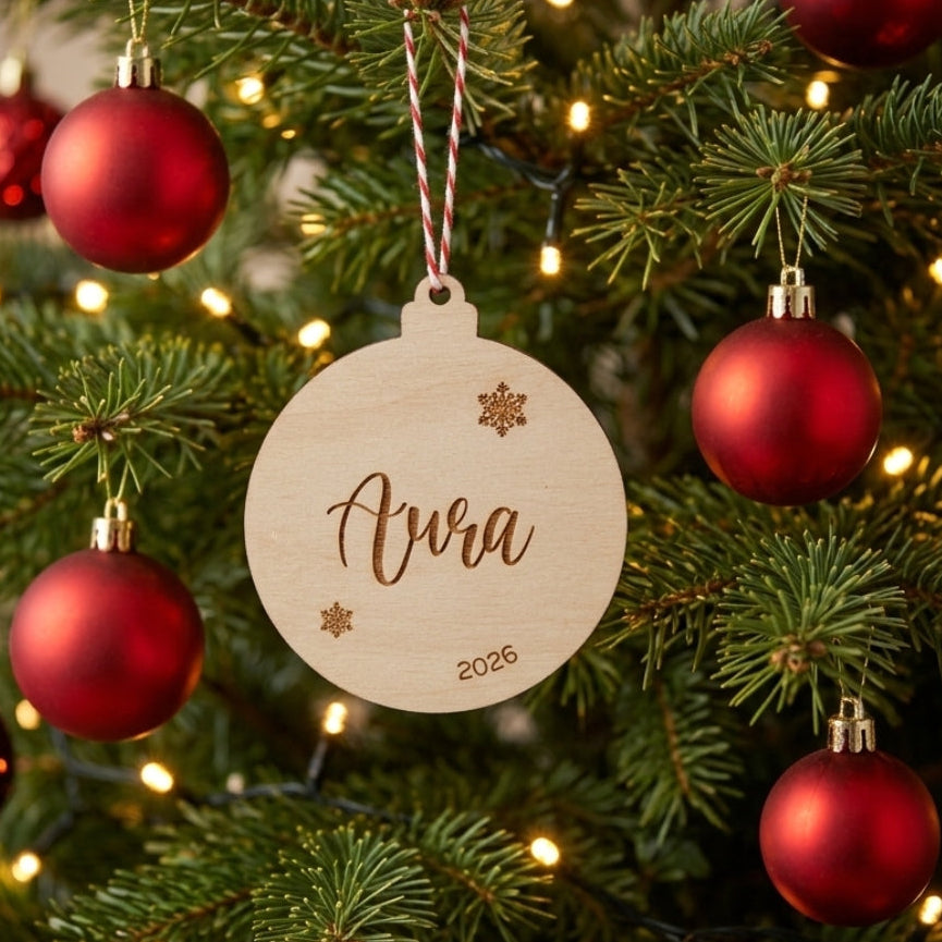 A personalized round wooden Christmas ornament engraved with the name 'Ava' and the year '2026', hanging on a decorated Christmas tree with red ornaments and warm lights.