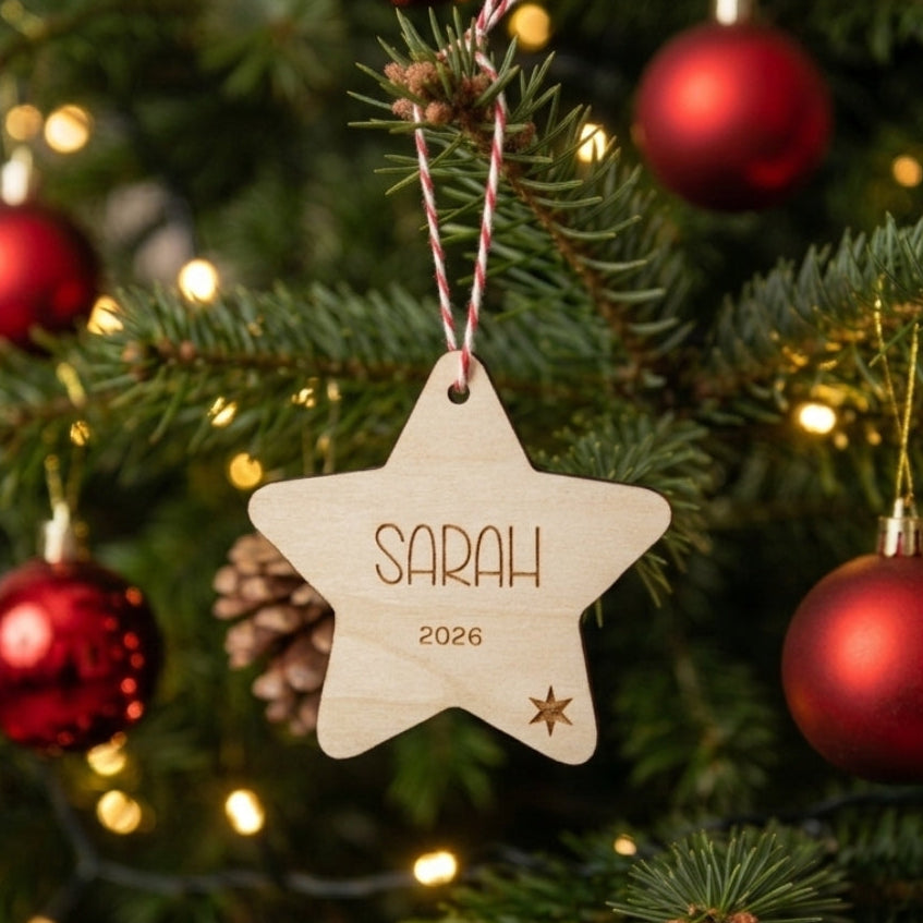 A personalized wooden star Christmas ornament hanging on a decorated Christmas tree. The natural wood star is engraved with the name 'SARAH', the year '2026', and a small star at the bottom, suspended by a red and white striped string.