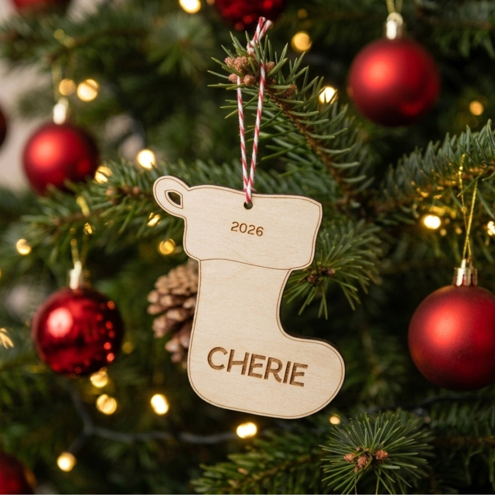 A personalized wooden Christmas stocking ornament hanging on a decorated Christmas tree. The natural wood stocking is engraved with the year '2026' and the name 'CHERIE', suspended by a red and white striped string.