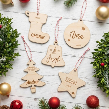 Six personalized wooden Christmas ornaments in stocking, bell, tree, star, and round shapes, each engraved with a name and the year 2026, hanging by red-and-white striped strings on a white wood surface surrounded by greenery and gold and red ornaments.