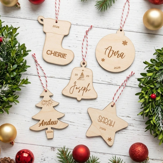 Six personalized wooden Christmas ornaments in stocking, bell, tree, star, and round shapes, each engraved with a name and the year 2026, hanging by red-and-white striped strings on a white wood surface surrounded by greenery and gold and red ornaments.