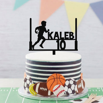 A custom black sports cake topper featuring a running football player silhouette, the name 'KALEB', and the number '10', displayed on top of a decorated birthday cake with football-themed decorations.