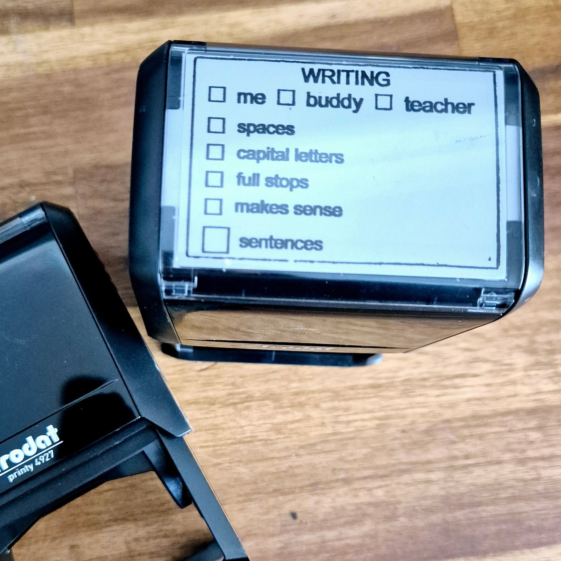 Trodat Printy 4927 Writing Feedback Stamp with customizable text plate and pre-designed checkboxes for writing elements like spaces, capital letters, full stops, makes sense, sentences. The stamp is self-inking and has a transparent positioning window.