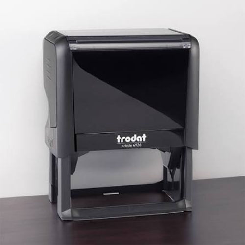A Trodat self-inking stamp, model printy 4926, in black plastic, shown standing on a dark wooden surface against a plain white wall.