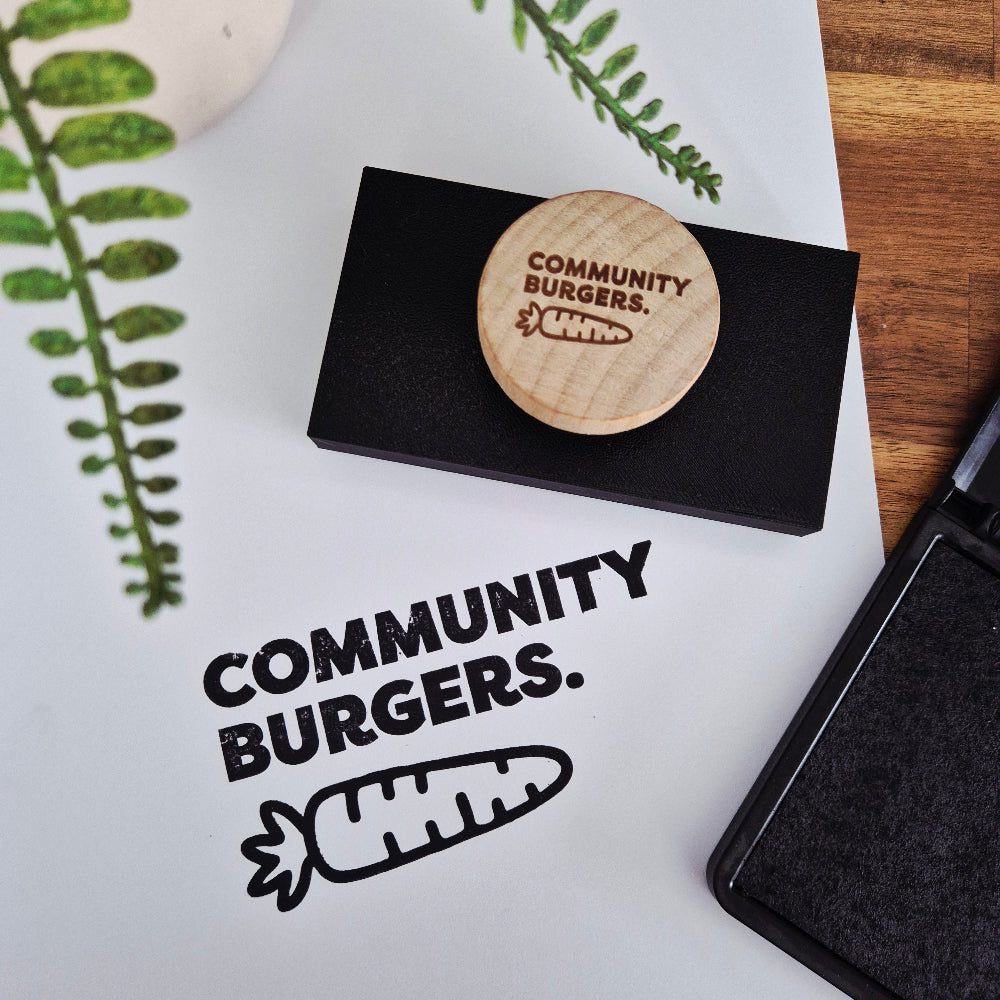 Wooden stamp with 'Community Burgers' branding on a white surface with a stamp and leaves.