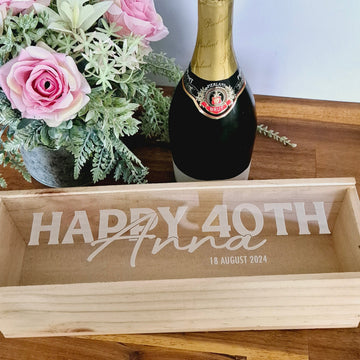 A clear acrylic bottle box with 'Happy 40th' engraved on it, displayed with a bottle and flowers.